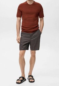 Man in een roestkleurig shirt met korte mouwen, grijze knielange shorts en zwarte sandalen met dubbele bandjes, staand met één hand in zijn zak.