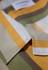 Tela a rayas en amarillo, verde, beige y marrón, con un borde de cuello visible y detalles de costura; presenta una etiqueta de tela en el fondo.