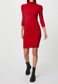 Robe rouge côtelée avec un col haut, un buste ajusté et des manches trois-quarts. Présente des détails de rayures verticales et une silhouette moulante.