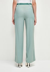 Jasnozielone spodnie o szerokich nogawkach z gładką strukturą, wysokim stanem i detalem w postaci tylnej kieszeni, zestawione z kremowymi szpilkami.