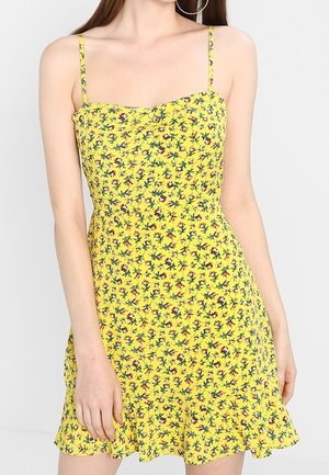 Robe fleurie jaune avec fines bretelles, corsage ajusté et ourlet évasé, ornée de petits motifs verts et rouges.