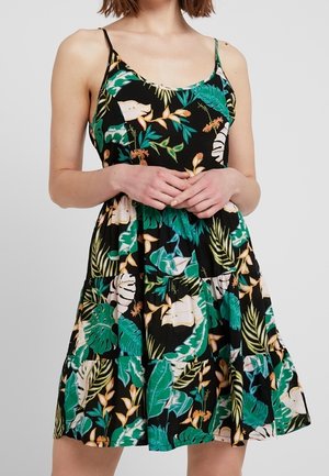 Frau trägt ein schwarzes ärmelloses Kleid mit grünen und beigen tropischen Blatt- und Blumenmustern, die Hände sanft an der Taille verschränkt.