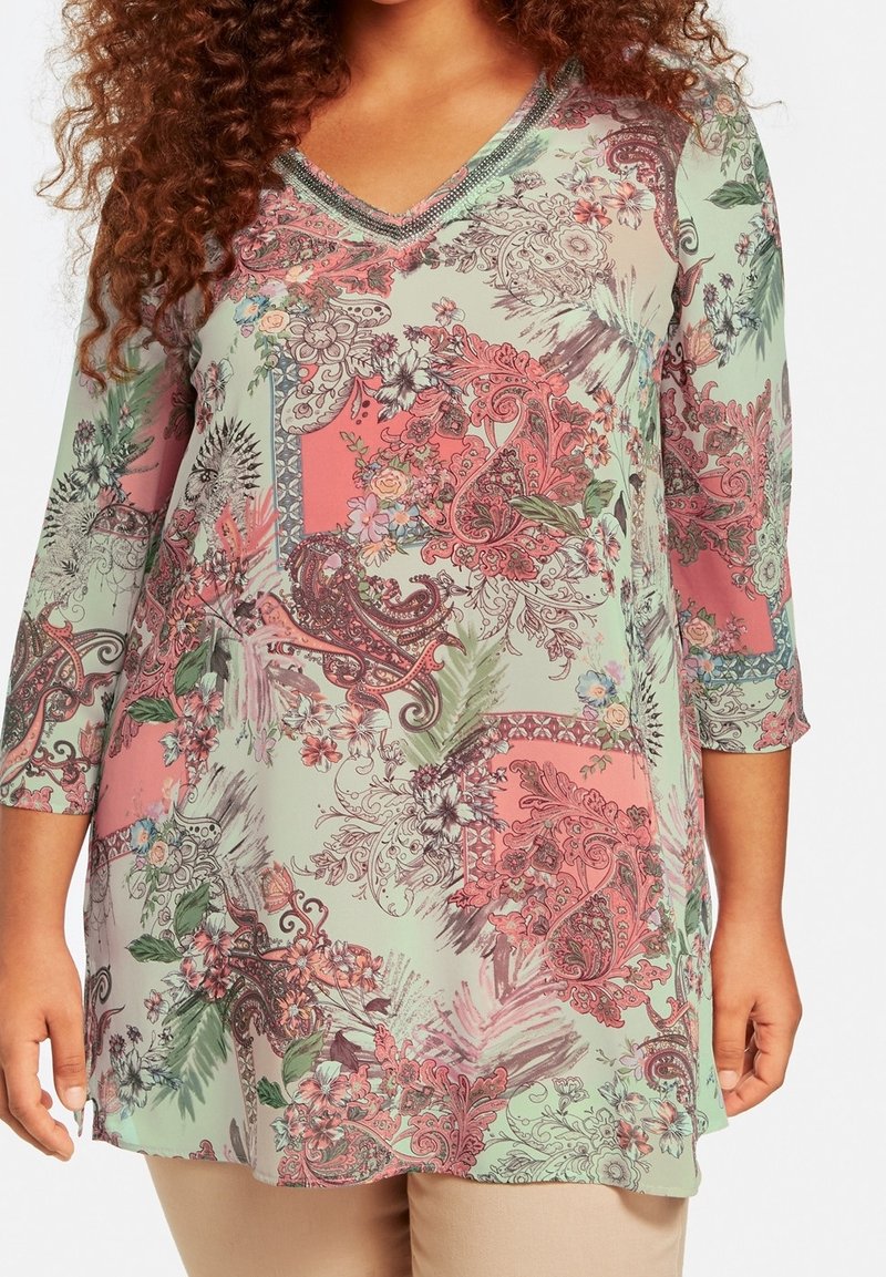 Femme portant une tunique à motifs floral paisley vert pastel et rose, manches trois-quarts et col en V.