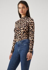 Žena u dugom rukavu s uzorkom leoparda i plavim trapericama, stoji nasuprot jednostavne svijetle pozadine, okrenuta blago lijevo.