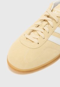 Svetlo rumeni semiš športni copati z belim dizajnom treh črt, okroglo konico in teksturirano gumijasto podplat, z vezalnim zapiranjem in gladkim šivanjem.