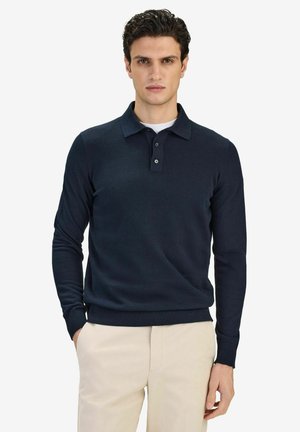 Man med mörkt lockigt hår, klädd i en marinblå långärmad polotröja och beige byxor, stående med ena handen i fickan.