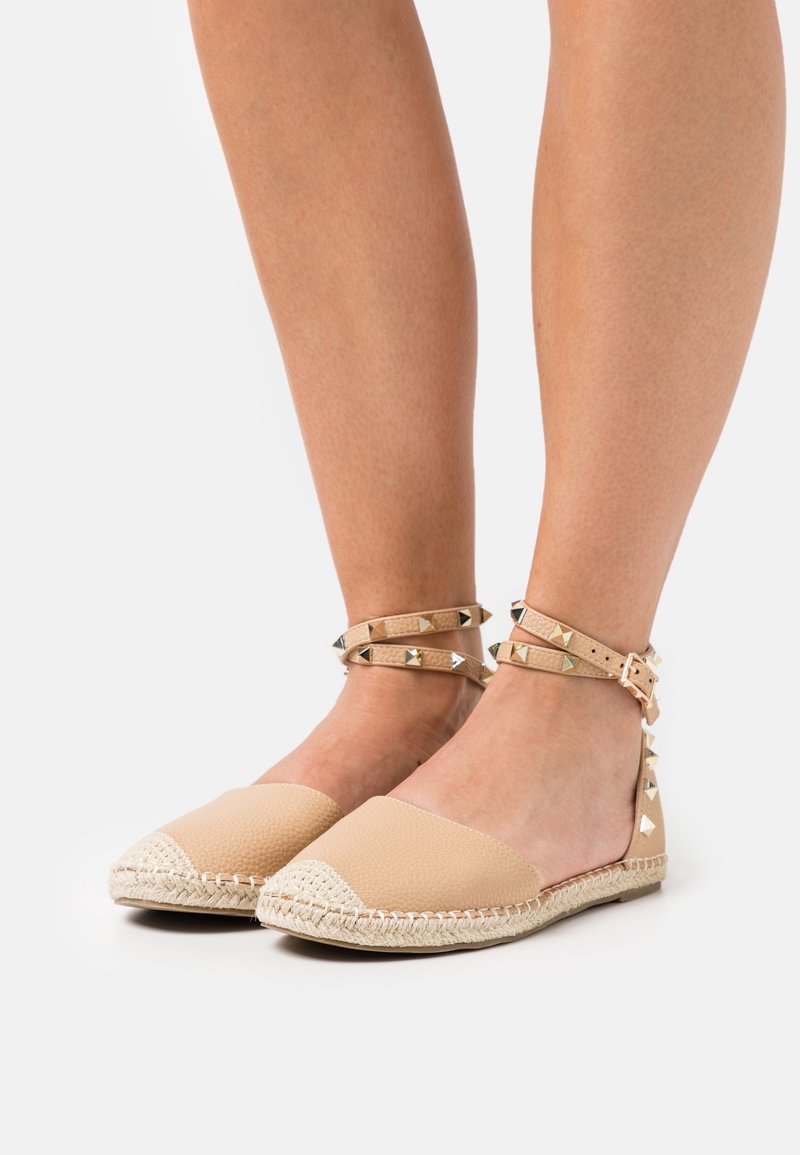 Jambes portant des espadrilles plates beige avec des brides de cheville cloutées et des bouts tissés, présentées sur un fond blanc.