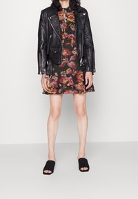 Musta nahkainen moottoripyöräilytakki tumman kukkamekon päällä, jossa on röyhelöitä. Mallilla on päällään mustat avonaiset sandaalit. Yksinkertainen harmaa tausta.