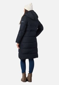 Lang, mørkeblå dunjakke med hætte, der har horisontal quiltning, sidelommer med lynlås, og som bæres med blå jeans og brune støvler.