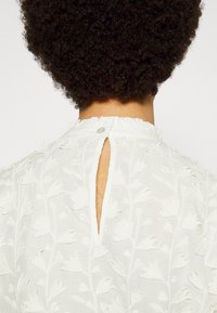Blouse blanche brodée avec des motifs floraux, une ouverture en forme de goutte dans le dos, et un col à volants avec une fermeture par bouton.