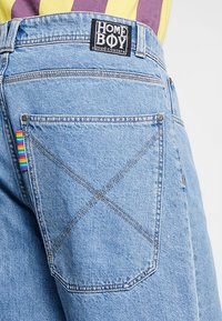 Hellblaue Jeans aus Denim mit einer großen Gesäßtasche mit X-Stich-Muster, einem schwarzen Markenlabel und einem mehrfarbigen gestreiften Etikett.