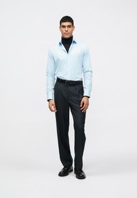 Camisa azul clara de botões por cima de um suéter de gola alta preto, combinada com calças escuras risca de giz e sapatos pretos. Design simples, ajuste sob medida.