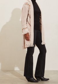 Long, beige cardigan with floral embroidery and fringe detail, worn over a ribbed black turtleneck, paired with black flared pants and ankle boots.
