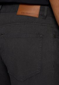 Zwarte denim broek met een gladde textuur, voorzien van een achterzak en een bruine leren patch met het label "TRUSSARDI" op de tailleband.