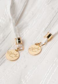 Hvidt stof med sølvstriber, der har to beige snore med guldfarvede metalender og runde, mærkede guldsymboler.