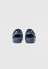 Tummansiniset synteettiset puutarhatossut, joissa on avoin takapää ja ilmanvaihtoaukot. Litteä, koostumukseltaan karkea pohja, jossa on "CROCS" -logo kohokuvioituna sivuilla.