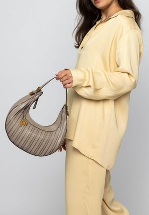 Beige pleated handbag with a curved shape, gold hardware, and a monogram accent. Worn with a light yellow silk blouse and pants.