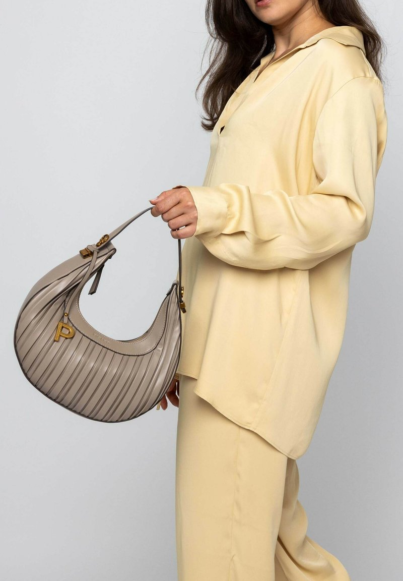 Beige geplooide handtas met een gebogen vorm, gouden hardware en een monogramaccent. Gekleed met een lichtgele zijden blouse en broeken.