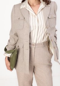 Blazer beige à chevrons avec boutons dorés, associé à une chemise crème à rayures. Tenant une petite pochette verte. Coupe ajustée, tissu texturé.