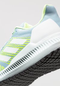 Scarpa sportiva azzurro chiaro con tomaia in rete, dettagli verdi e strisce bianche. Presenta una suola testurizzata e un tallone imbottito per il supporto.