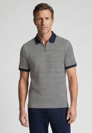 Man draagt een navy- en witgestreept polo met korte mouwen, een navy kraag en manchetten, gecombineerd met navy broeken, staand tegen een effen achtergrond.