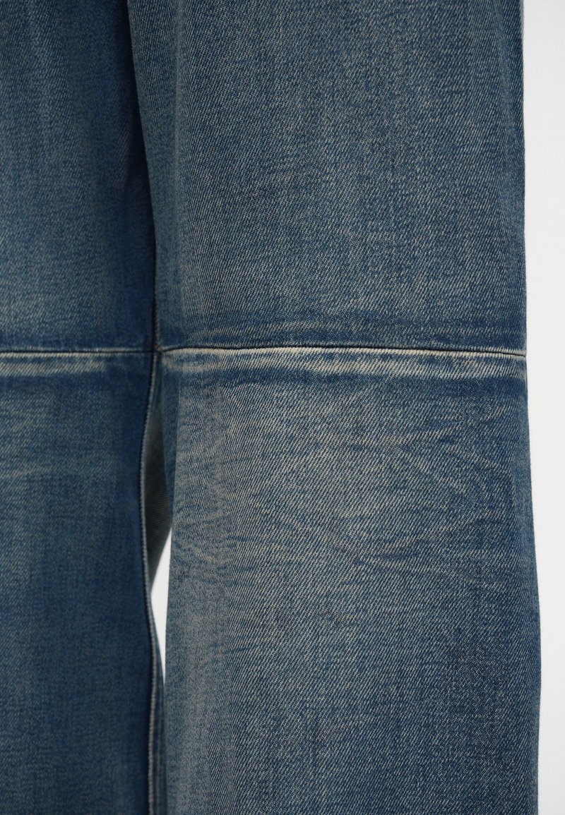 Jean en denim de couleur bleu moyen, présentant une texture lisse, des coutures visibles et un détail légèrement décoloré au niveau du buste.