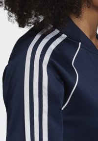 Osoba s kudrnatými vlasy, která má na sobě námořnickou modrou bundu se třemi bílými proužky probíhajícími po rameni a bílými detaily.