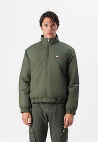 Olivengrønn zip-up jakke med høy krage, elastiske mansjetter og hem. Har en logo-patch i rødt, hvitt og blått på brystet.