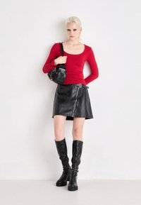 Haut ajusté à manches longues rouge, mini-jupe noire en simili cuir avec fermeture éclair sur le côté, bottes à plateforme noires jusqu'aux genoux, et un petit sac à main noir.