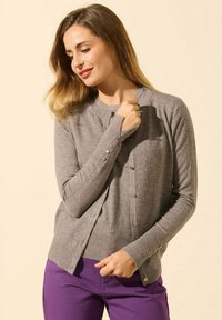 Gilet en tricot gris avec un col rond et des boutons sur le devant et les poignets, associé à un pantalon violet. Texture lisse.