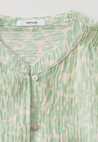 Gros plan d'un chemisier à motif vert clair et beige avec encolure ronde, fermeture à boutons et détail de tissu froncé à l'épaule.