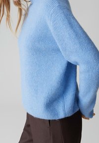 Femme portant un pull tricoté pelucheux bleu clair et un pantalon marron foncé, vue de profil sur un fond uni.