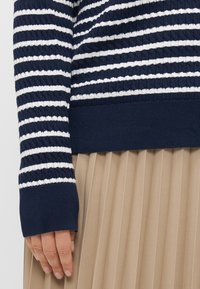 Navy and white striped knit sweater with textured fabric, featuring a ribbed cuff. Worn over a beige, pleated skirt.
