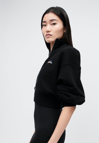 Čierny skrátený sweatshirt s vysokým golierom a zipsom. Má pufované rukávy a malé logo na hrudi. Mäkká textúra.