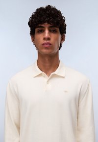 Crème kleurige lange mouwen polo shirt met kraag, voorzien van twee knopen en een klein geborduurd logo op de linkerkant van de borst. Gladde textuur.