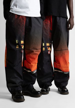 Široke crne hlače s apstraktnim crvenim i narančastim uzorkom, koje imaju glatku teksturu i malu oznaku "WASTED" blizu ruba.