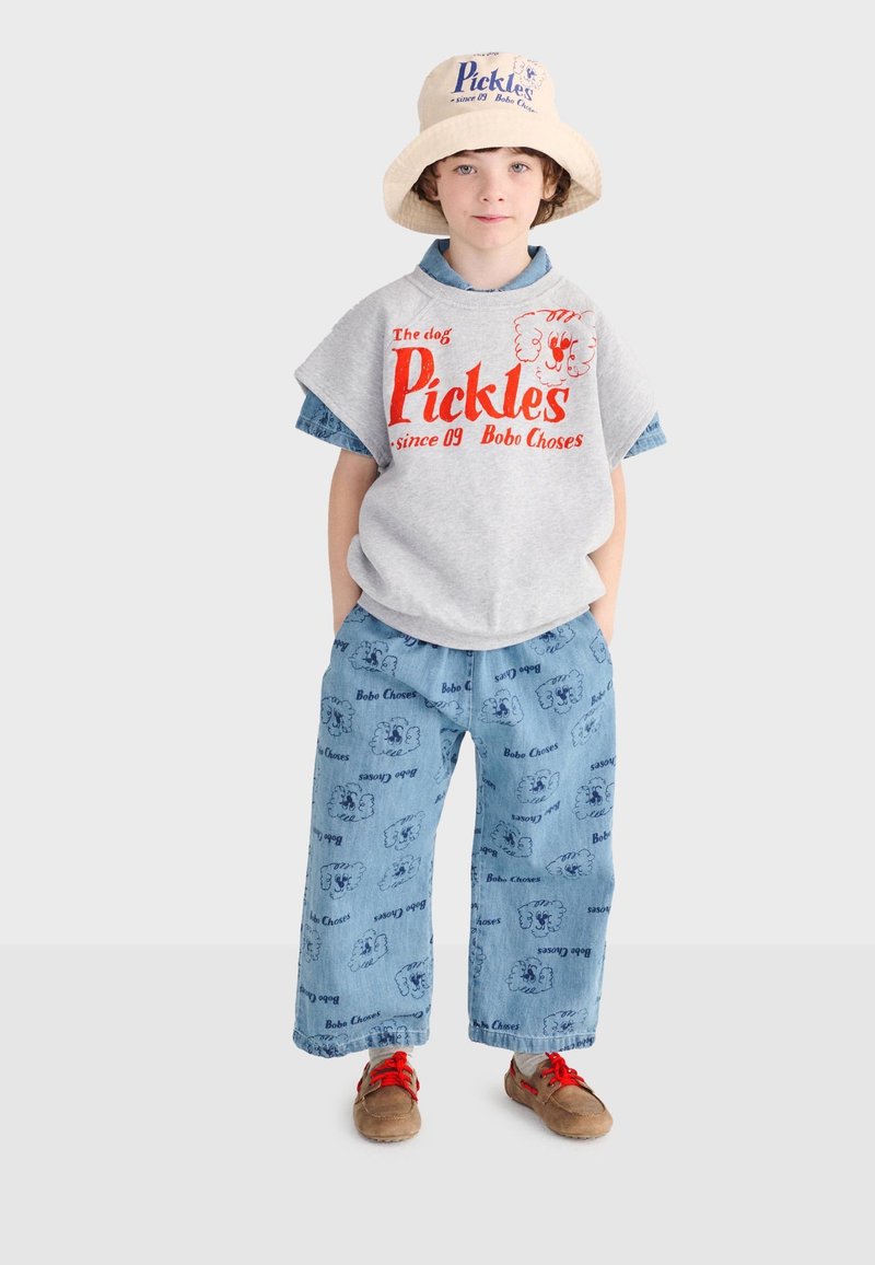 Enfant portant un chapeau seau beige, un débardeur gris avec le texte rouge "Pickles", un pantalon large bleu à motifs de chiens, et des chaussures marron avec des lacets rouges.