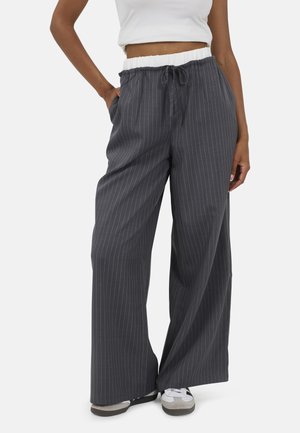Persoon draagt donkergrijze wijde pinstripe broek met een witte elastische tailleband en witte sneakers, staand met één hand in de zak.