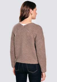 Brauner Strickpullover mit V-Ausschnitt hinten, geripptem Saum und fallenden Schultern. Strukturierter Garn sorgt für ein weiches Aussehen. Über dunklen Jeans getragen.