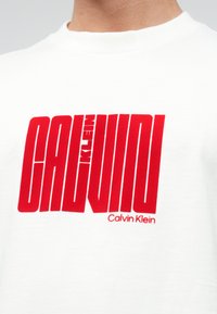 White cotton t-shirt featuring prominent red graphic text "CALVIN KLEIN" across the chest and logo detail below in a bold, modern font.