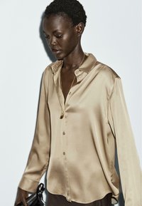Camicia beige in satin con bottoni, colletto, maniche lunghe e orlo arrotondato. Presenta una leggera lucentezza e una collana con pendente.