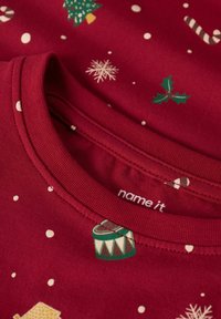 Tessuto natalizio rosso con vischio verde, fiocchi di neve bianchi, bastoncini di zucchero e motivi di tamburo attorno a un colletto rotondo.