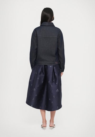 Woman with long dark hair wearing a dark denim jacket, navy floral midi skirt, and white heeled sandals, standing facing away on a white background.