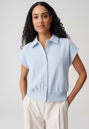 Vrouw met lang, krullend haar die een lichtblauw kort mouwens overhemd en crèmekleurige broek draagt, staand met de handen in de zakken.