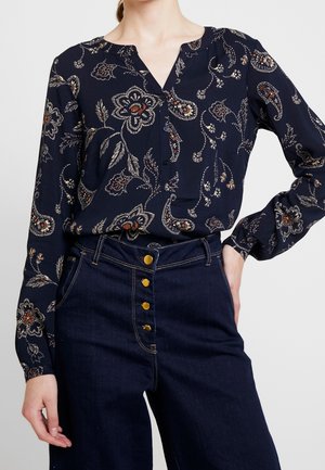 Kvinde iført navyblå blomstret langærmet bluse, stoppet ned i højtsiddende mørke denimjeans med synlige guldknapper.