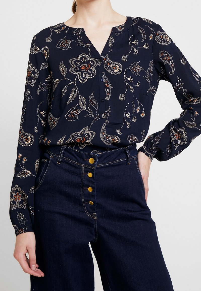 Femme portant un chemisier bleu marine à fleurs à manches longues, rentré dans un jean taille haute en denim foncé avec des boutons dorés visibles.