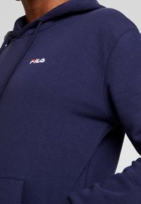Navy blue hoodie with a kangaroo pocket, featuring a small white and red embroidered Fila logo on the chest, made of soft cotton material.