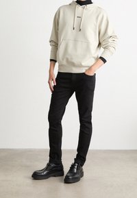 Beige bavlněná hoodie s tkaničkami a přední kapsou, spárovaná s černými slim fit džíny a černými koženými botami s tlustou podrážkou.