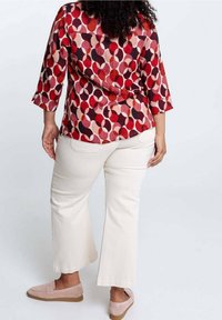 Donna che indossa una blusa a fantasia rossa, rosa e nera, pantaloni bianchi a vita corta e scarpe slip-on rosa chiaro, in piedi su uno sfondo bianco.