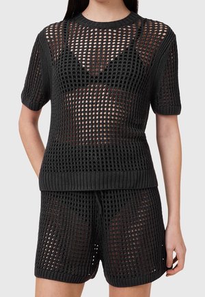 Femme portant un haut court à manches courtes en maille noire tricotée et un short assorti avec des sous-vêtements noirs visibles dessous.